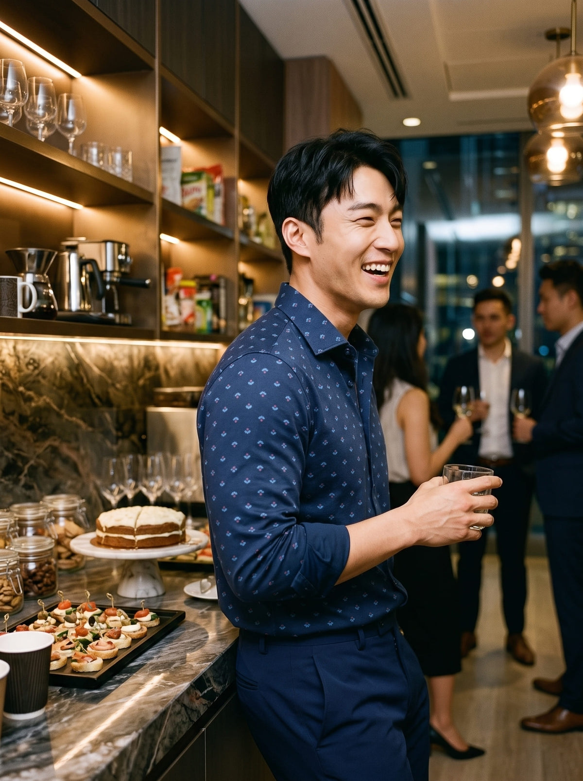 A man in a navy blue patterned button-down shirt and matching trousers stands in a modern bar or lounge setting, smiling and holding a glass. Shelves with glasses and coffee equipment are visible behind him, along with other people in the background.