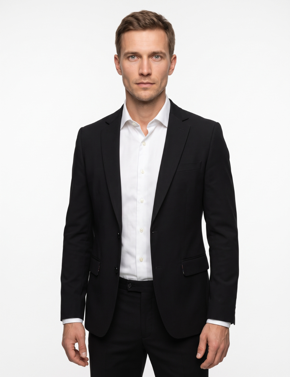 A man wearing a black blazer over a white dress shirt and black trousers, standing against a plain light gray studio background. #Color_Black