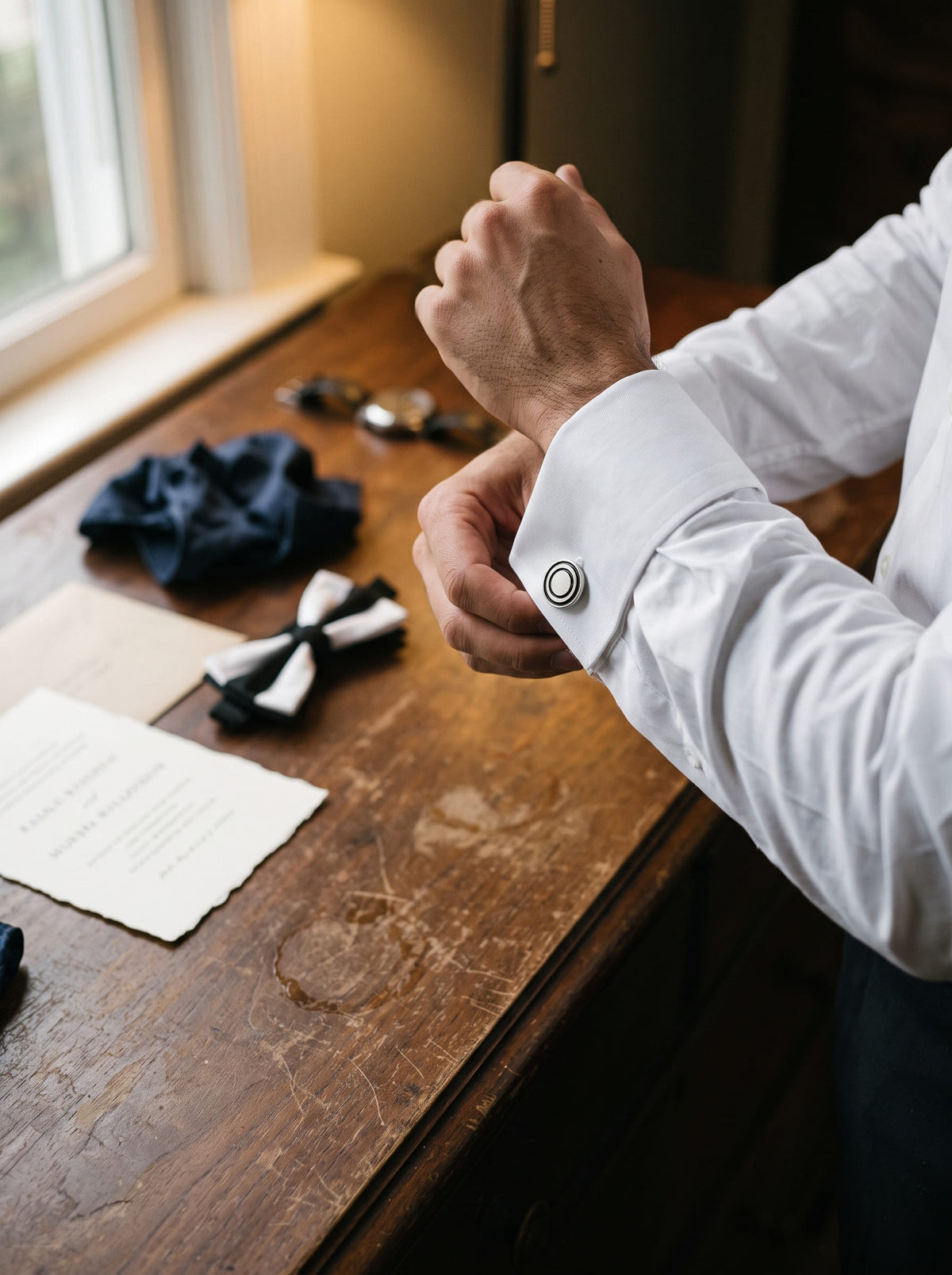 A close-up lifestyle shot of a man's hands adjusting a silver cufflink on a white dress shirt sleeve, set against a rustic wooden table with a navy blue tie and a bow tie.